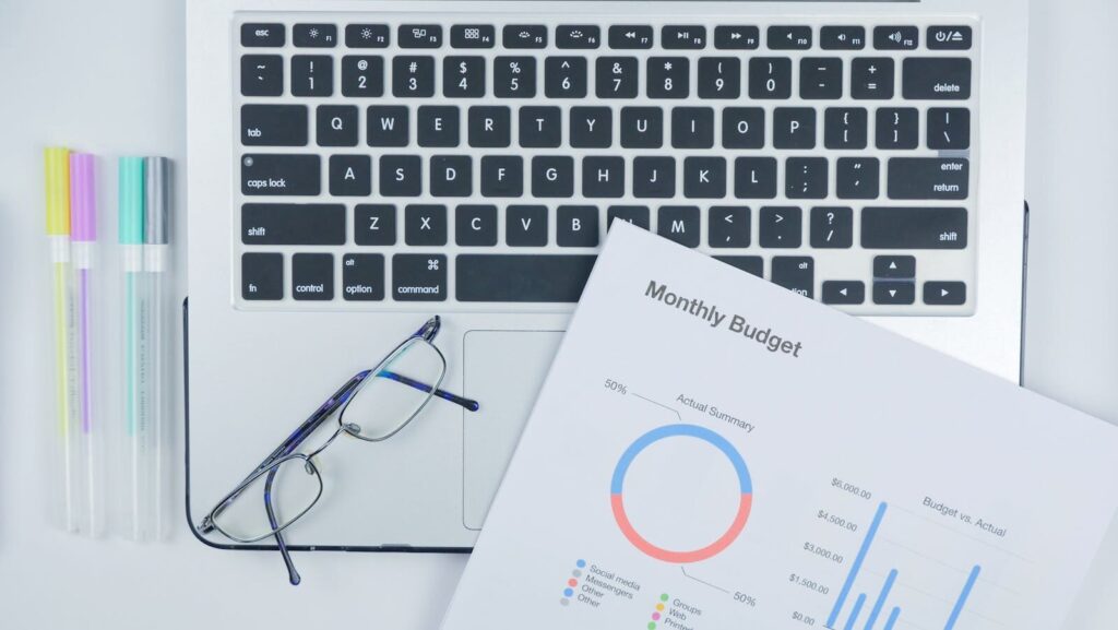 Aerial shot of a modern workspace with a laptop, budget report, and eyeglasses on a desk.
